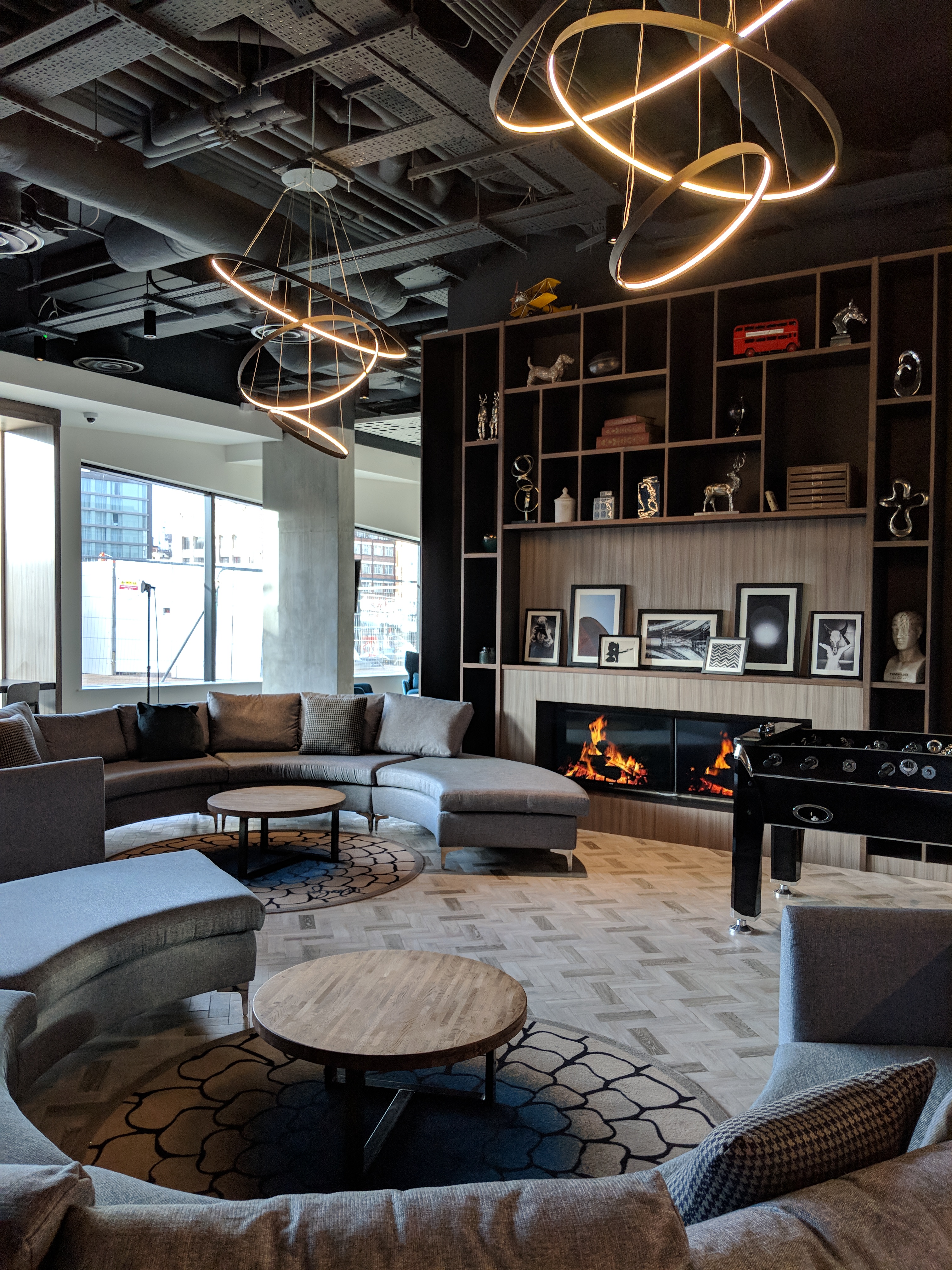 Modern lounge featuring circular gray sofas, a foosball table, and two coffee tables. A fireplace with decorative shelves above enhances the cozy ambiance. Large windows provide natural light, and circular ceiling lights add a stylish touch.